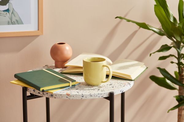 Still life of pale yellow mug of coffee with notebook and open book on round marble table.