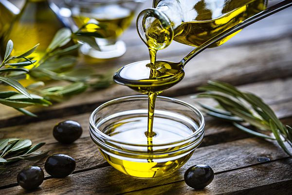 Olive oil being drizzled into a cup on a wood table with herbs and olives.