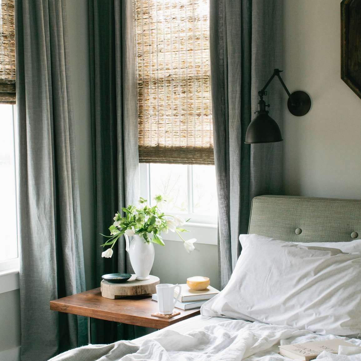 两扇窗户旁边的床上有凌乱的床单，还有一个放着一盆植物和一杯咖啡的床头柜