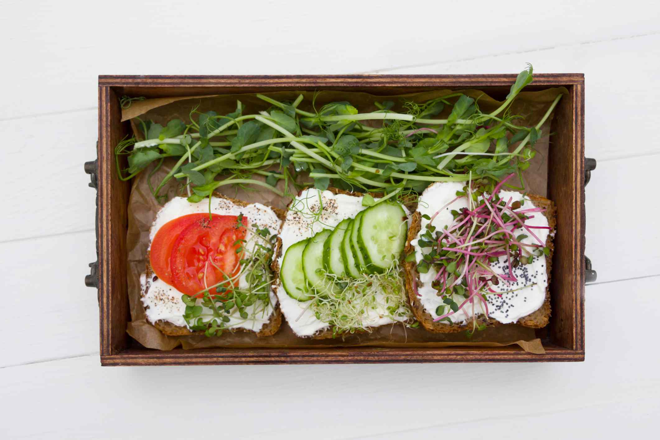 木制的盒子里放着三片吐司，上面有奶油芝士，西红柿，黄瓜和旁边有豌豆芽的绿色蔬菜