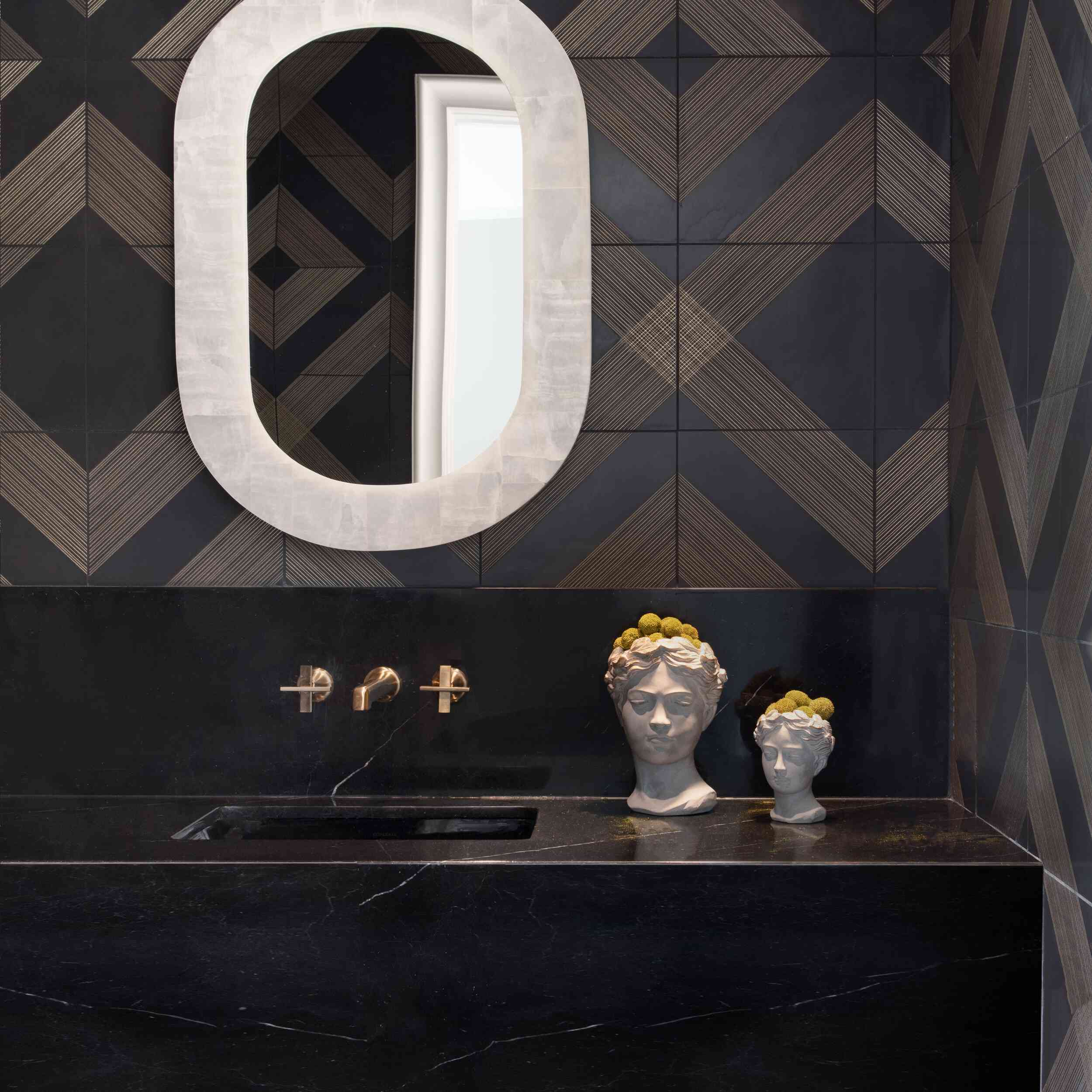 一间暗色的化妆室，墙上有金色和黑色的图案，一个黑色的水槽，一面白色的镜子